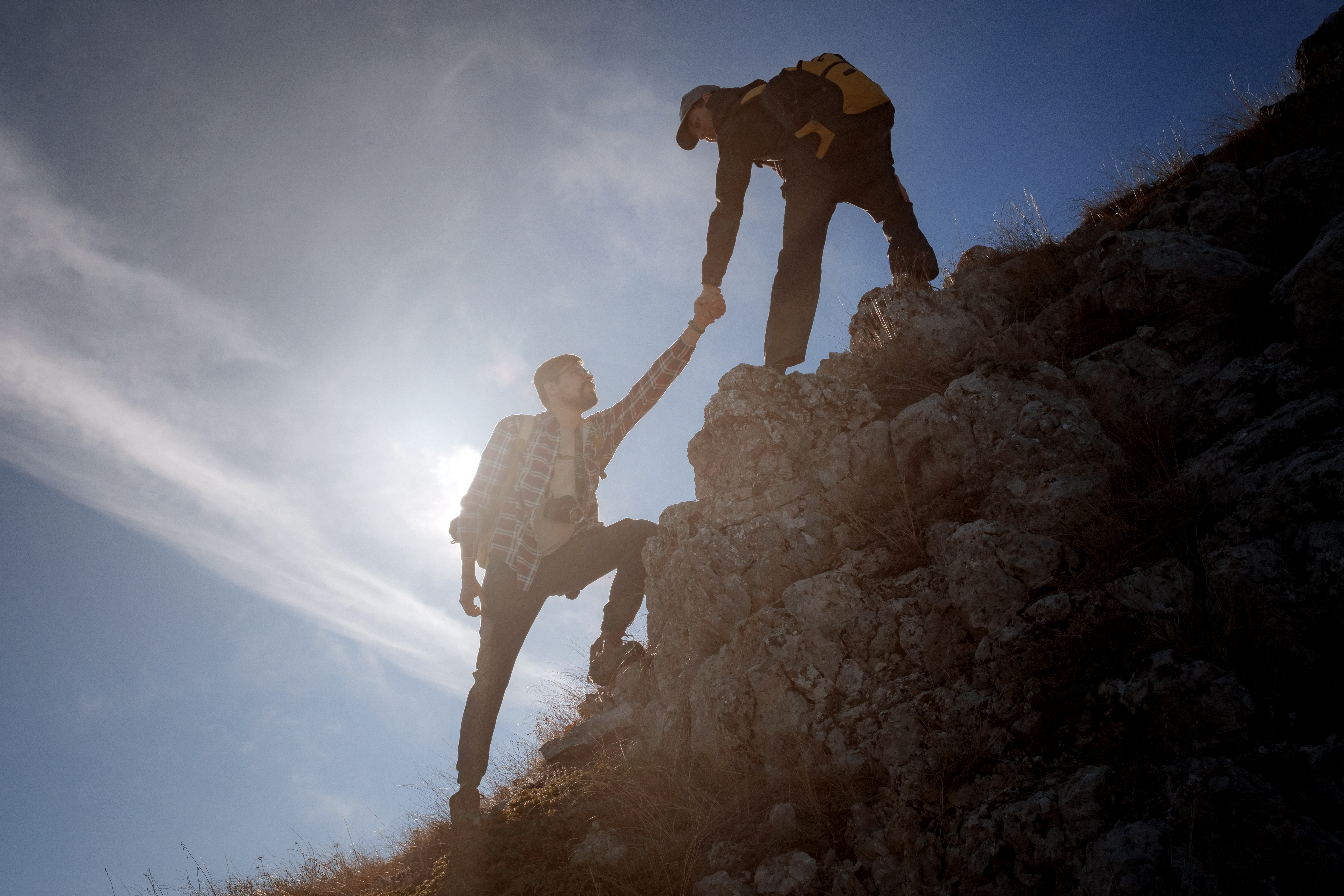 Silhouettes of two people climbing mountains and helping against the blue sky.